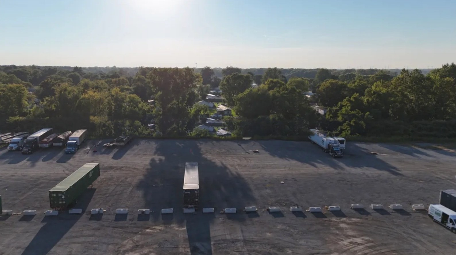Low aerial view across the gravel lot at sunrise, with trailers and a tractor parked along the tree line